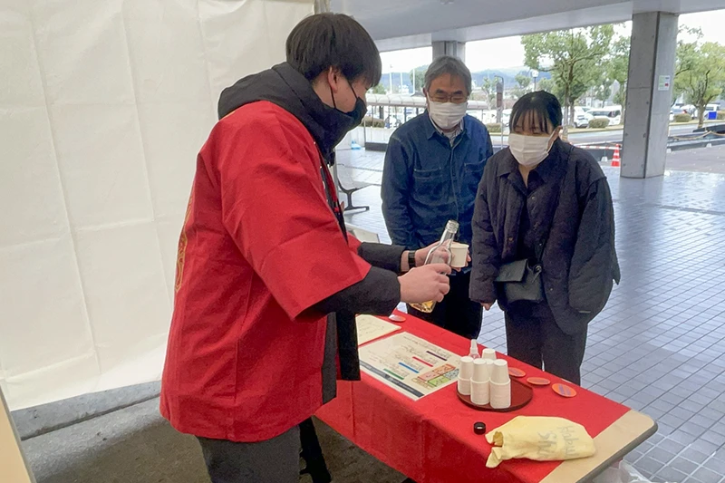 人吉カルチャーパレスの会場で開催された球磨焼酎の試飲会の様子。法被を着たスタッフが、来場した男女に球磨焼酎を注いでいる。
