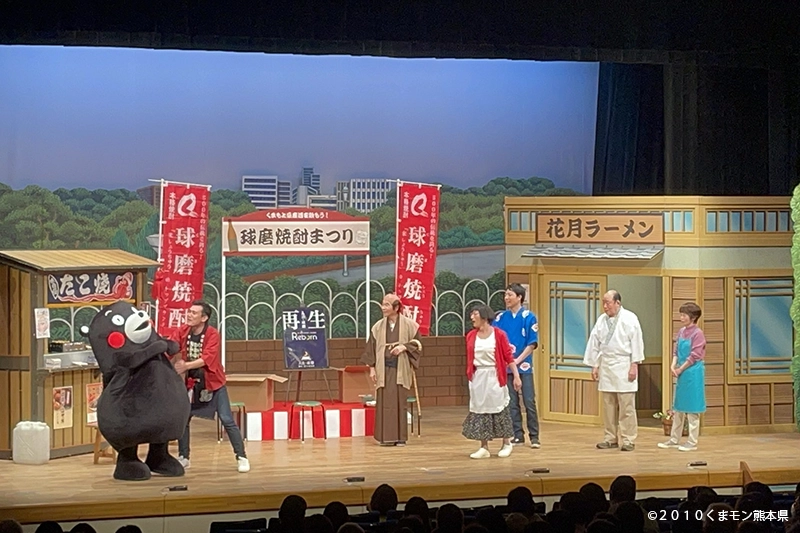 吉本新喜劇人吉特別公演の舞台の様子。花月ラーメンのセットの前で、くまモンと出演者たちがコミカルな演技を披露している。背景には「球磨焼酎まつり」ののぼりが見える。